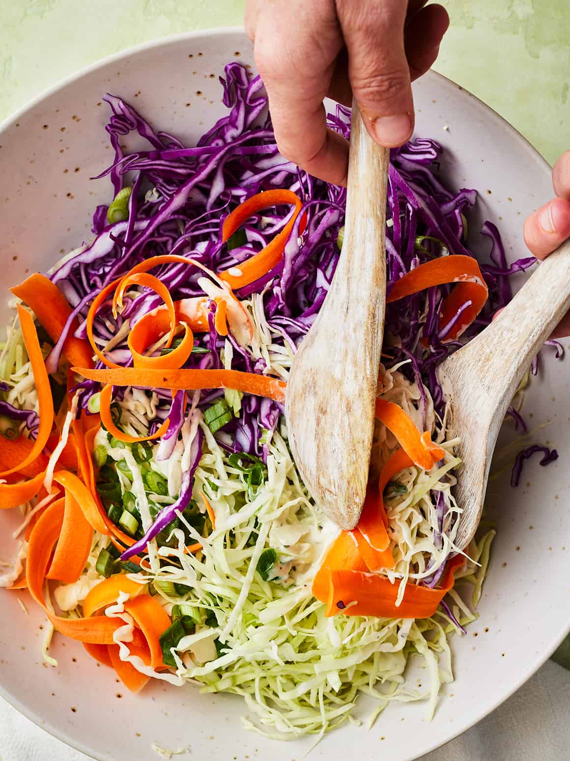 Hands tossing together shredded cabbage, carrots, and scallions with wooden spoons